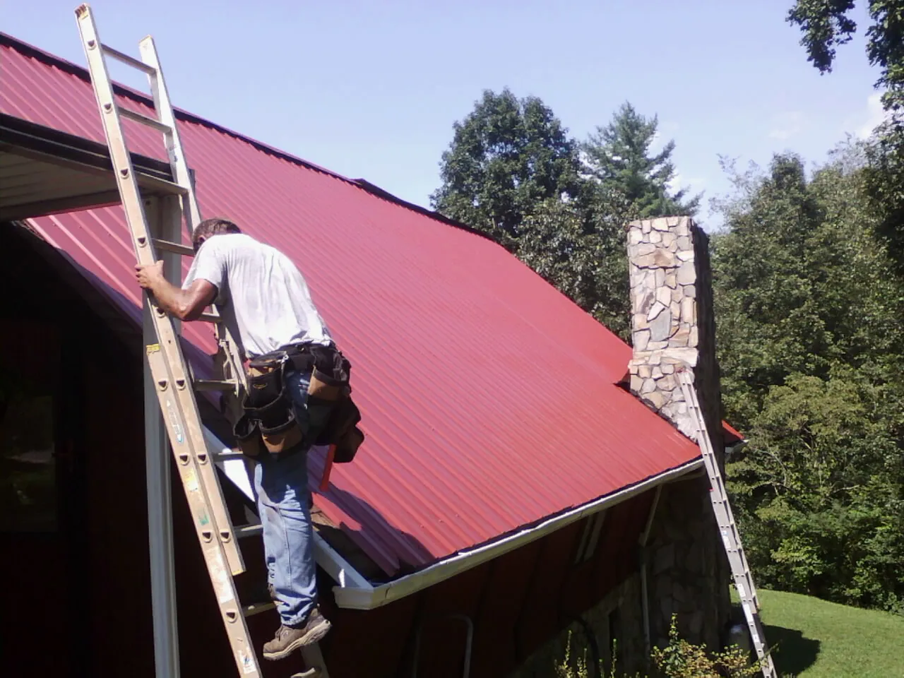 Roofer on a ladder beside a red metal roof and stone chimney — Soffit & Fascia Repair by Roofing Brothers in Lake